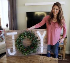 a woman standing next to a wooden joy sign with a wreath on it and the word joy written in large letters