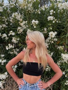 a blonde woman standing in front of some white flowers and bushes with her hands on her hips