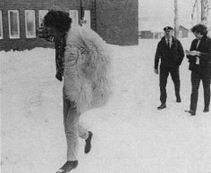 two men are standing in the snow with one man wearing a fur coat and another is looking at his cell phone