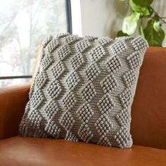 a gray knitted pillow sitting on top of a brown leather couch next to a window