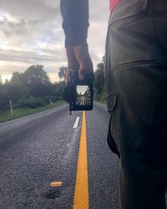 a person holding a camera while standing on the side of an empty road with trees in the background