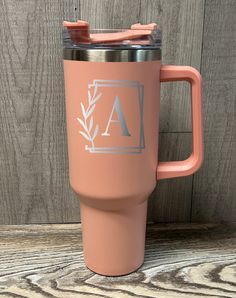 a pink travel mug with the letter a on it sitting next to a wooden wall