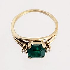 "This magnificent vintage ring boasts a lovely emerald and two diamonds set in 18K yellow gold. The emerald has a rich bluish-green color and weighs in at 1 1/4 carats. An accent diamond is set on each side of the emerald, adding elegance and sparkle. The ring has triple branched shoulders that add interest beyond the classic shank. Luxurious! Vintage: 1990s Size: 6 Materials: 18K Yellow Gold, Emerald, Diamonds Marked: 18K Emerald: 7 mm x 7mm x 4.4mm, approx. 1.25 ct. 2 Diamonds: brilliant cut, Asscher Cut Emerald Ring With Diamond Accents, 14k Gold Green Diamond Princess Cut Ring, Green Princess Cut Diamond Ring In 14k Gold, Princess Cut Green Diamond Ring In 14k Gold, Timeless Gold Diamond Ring With Emerald, Emerald Cut Green Diamond Ring With Diamond Accents, Emerald Cut Green Diamond Ring With Accents, Green Asscher Cut Ring With Diamond Accents, Vintage Green Diamond Ring With Center Stone