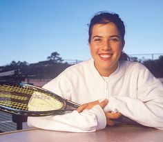 a woman sitting at a table with a tennis racket in her hand and smiling