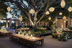 an outdoor dining area with tables, chairs and lanterns hanging from the trees in front of it