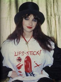 a woman with long hair wearing a black hat and lipstick stick t - shirt in front of a curtain