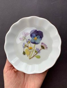 a hand holding a white bowl with purple and yellow flowers on the inside of it