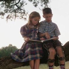 two children are sitting on a tree branch and reading a book in front of them