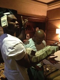 two men standing in a hotel room with money wrapped around their heads and one man holding up his hand