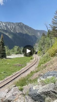 a train traveling through a lush green countryside next to trees and mountains on a sunny day