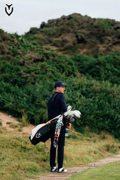 a man standing on top of a lush green hillside holding a golf club and bag