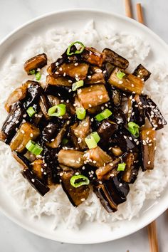 a white plate topped with stir fried eggplant and rice next to chopsticks