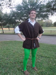 a man wearing green tights and a brown vest standing in the grass with his hands on his hips