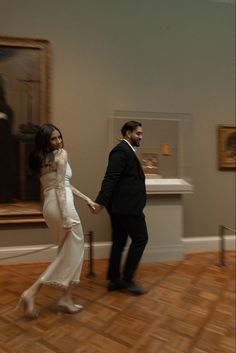 a man and woman in formal wear walking through a room with paintings on the wall