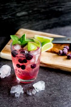 a drink with blueberries and lime on the rocks next to a cutting board full of ice cubes