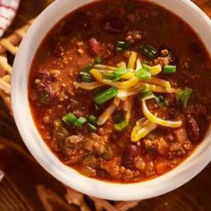 a white bowl filled with chili, cheese and bread on top of a wooden table