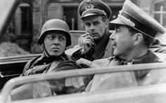 three men in uniforms are sitting in a car talking on the phone while another man sits behind them