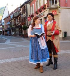 two people dressed up in costumes standing on a street corner with buildings and shops behind them