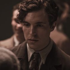 a man with curly hair wearing a suit and tie looking at the camera while standing in front of other men