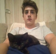 a man sitting on a couch holding a black cat in his lap and looking at the camera