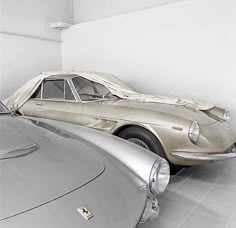 two old cars are parked in a room with white walls and tile flooring, one is silver and the other is beige