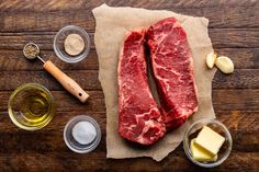 two raw steaks sitting on top of a wooden table next to spices and seasonings