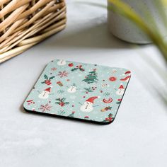 a small square coaster with snowmen and christmas trees on it next to a wicker basket