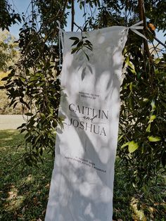 a sign that is hanging from a tree in the grass with leaves on it and some trees behind it