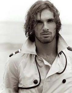 a man with long hair wearing a trench coat and standing on the beach in front of the ocean