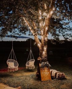 two hanging chairs next to a tree with string lights on it and some blankets underneath