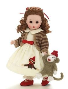 a doll is holding a stuffed animal and wearing a white dress with red trims