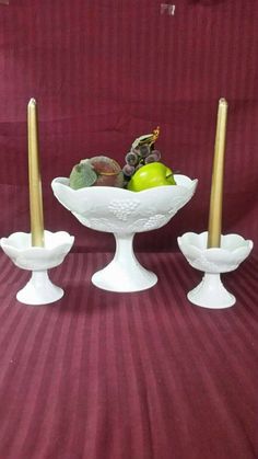 two white candlesticks are sitting in a bowl with fruit on it and one candle is next to them