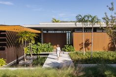 a woman walking down a walkway in front of a house with trees and plants on both sides