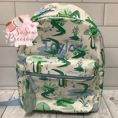 a white backpack with green and blue cactus print on the front, sitting on a wooden floor