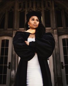 a woman wearing a graduation gown and holding her hands on her chest, standing in front of a building