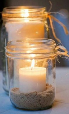 two lit candles are in a mason jar on a table with sand and twine