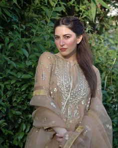 a woman standing in front of some bushes wearing a brown dress with gold trims