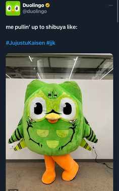 a green stuffed animal with big eyes and an orange beak standing in front of a white wall