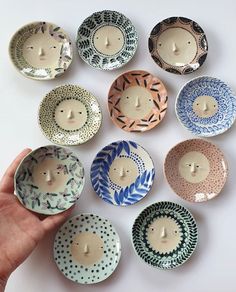 a hand is holding a small bowl with many designs on it and there are nine bowls in the shape of people's faces