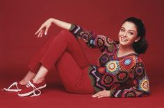 a woman is sitting on the floor wearing red pants and a colorful sweater, smiling at the camera