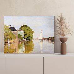 a painting on a shelf next to a vase with a plant in it and a house by the water