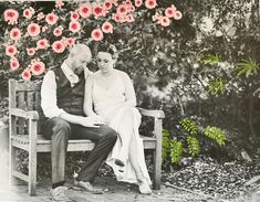 a man and woman sitting on a bench in front of some red flowers with one holding the other's hand