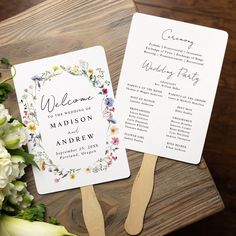 two wedding programs on top of a wooden table next to white flowers and greenery