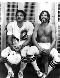 two men sitting next to each other with football helmets on their feet and one holding a ball