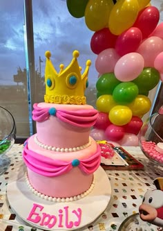 a pink cake with a crown on top and balloons in the background at a birthday party