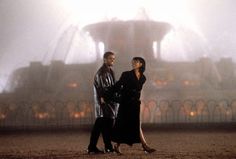 a man and woman walking in front of a fountain on a foggy day,