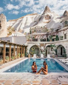 two women sitting on the ground in front of a swimming pool with mountains in the background