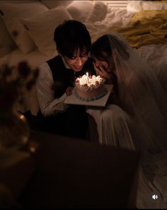 two people sitting on a bed looking at a cake with lit candles in the middle