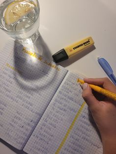 a person is writing on a notebook with a yellow pen and a glass of water