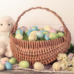 a teddy bear sitting next to a basket filled with painted easter eggs and flowers on a table